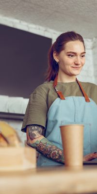 Female cafe worker in apron standing behind counter and working on modern notebook